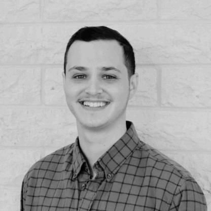 A person smiling in front of a brick wall, wearing a plaid shirt.