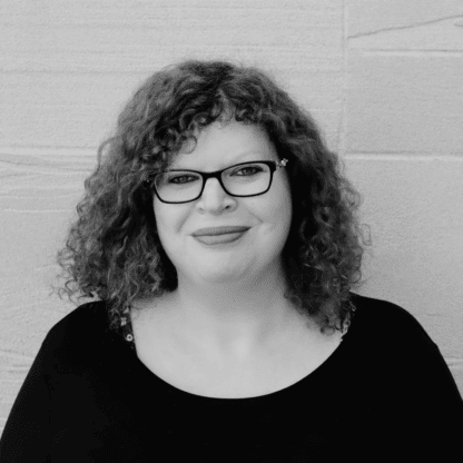 A person with curly hair and glasses smiling slightly, wearing a black top, stands in front of a textured wall. The image is in black and white.