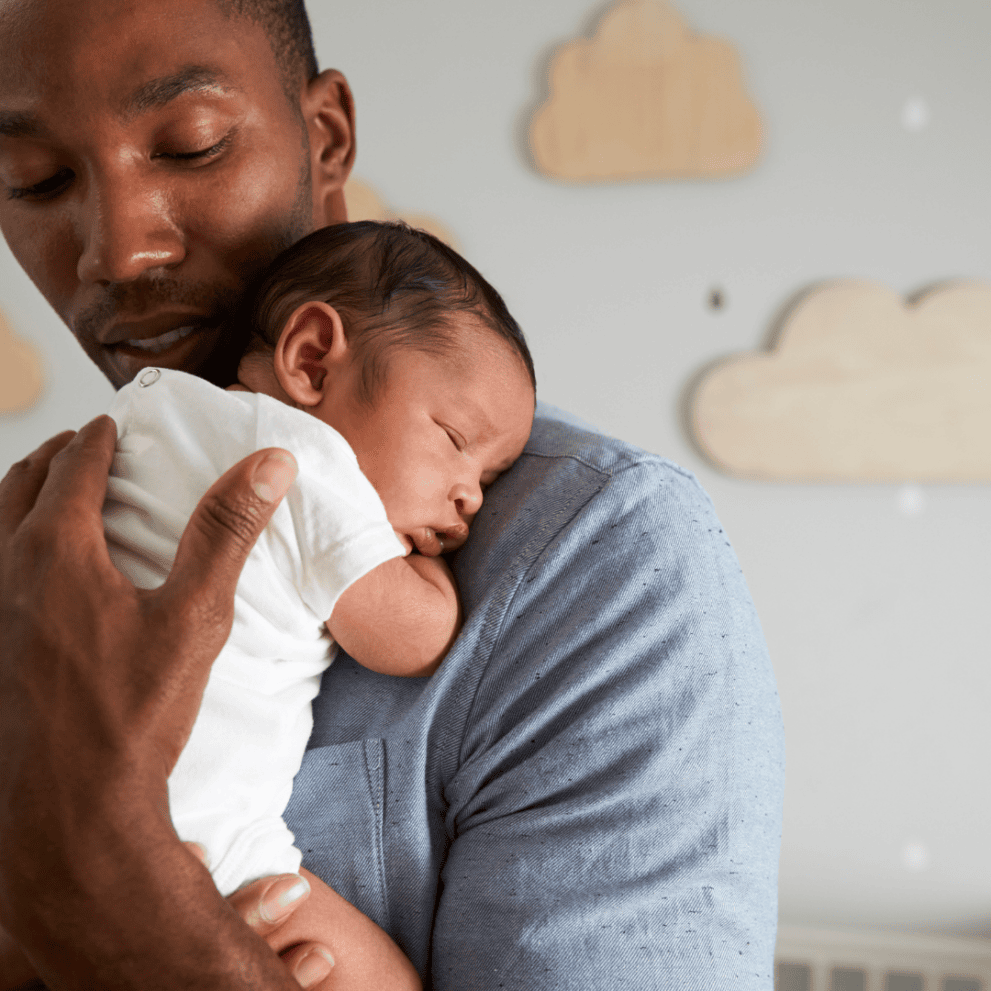 A man holds a sleeping baby closely on his shoulder in a room with wooden cloud decorations on the wall.