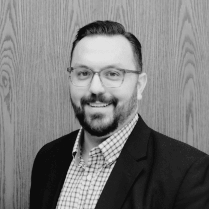 A man with glasses and a beard, wearing a checked shirt and blazer, stands smiling in front of a wooden panel background. The image is in black and white.