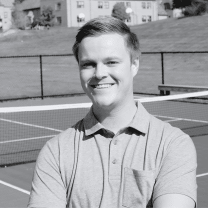 A smiling person stands on a tennis court in front of a net, with buildings and grassy hills in the background. The image is in black and white.