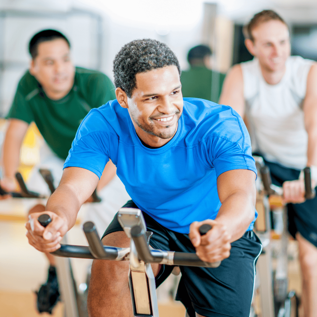 Three people are riding stationary bikes in a gym, smiling and wearing athletic clothing.