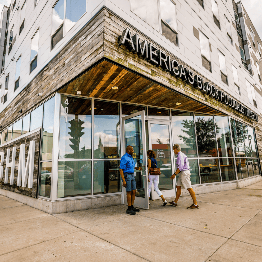 Three people standing outside the entrance of America's Black Holocaust Museum, a modern building with large windows and wood accents.