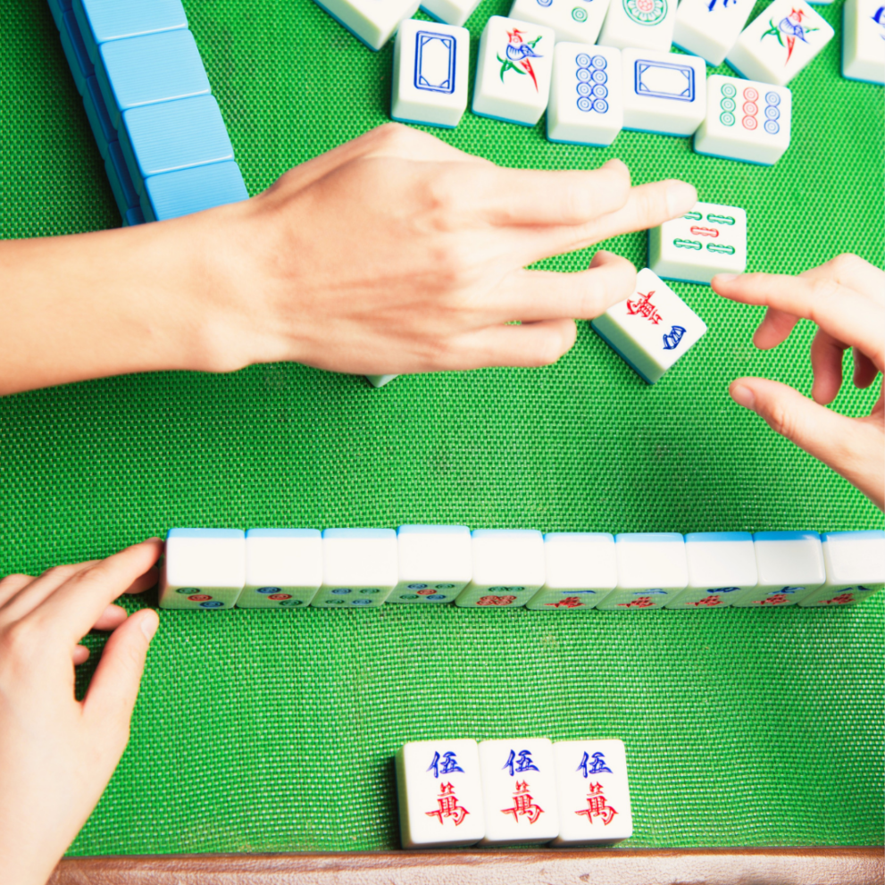 Hands arranging and playing Mahjong tiles on a green mat.