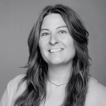 Smiling woman with long hair and a nose ring, wearing a light top, against a plain background. Black and white photo.