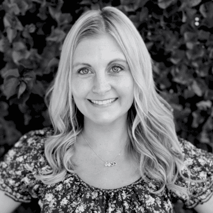 Woman with long blonde hair and a floral blouse smiling in front of leafy background. Black and white photo.